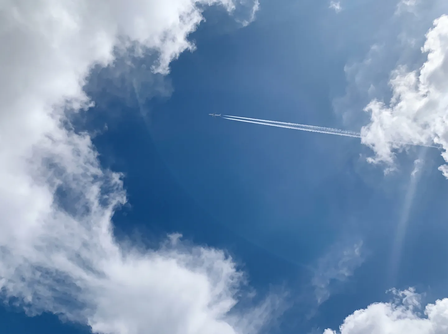 A passenger jet far up in a clear blue sky leaving a long white trail, seen from the ground through a gap in clouds